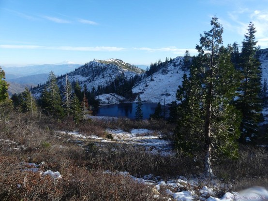 Lily Pad Lake from the Pacific Crest Trail. Mount Shasta is a faint giant peak in the distance.

A lake with ice around the edges sits in a bowl. The far side has a bit of snow, the near side has much less snow with bushes sticking up out of it, their leaves red-brown with the season. A few bright green conifers stick up in groups, one near to the left of the lake and others along a ridge edge behind the lake and one very near on the right of the lake. The ridge climbs and then drops away behind things with more ridges lining the horizon behind it. Above the furthest ridge line, a tall white peak, vanishing in the thin cloud of winter warming smoke, sticks up into the sky.