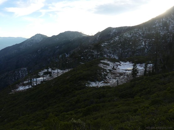 Kangaroo Springs along the Pacific Crest Trail.

Green brush cover a slope dropping to the left. In the near distance, two flats break out to the right of the slope, covered in a layer of snow. Small depressions are visible in the flats. Behind, a ridge line rises to a couple little peaks and drops. The sky is bright while the land is in shadow.
