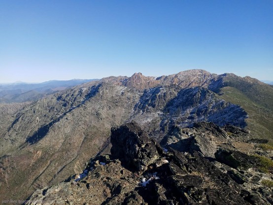 Near the top of Rattlesnake Mountain, looking east toward Red Buttes.

Looking back along a ridge. Dark rock rises along the ridge in the foreground. Green shrubs rise up the right side and grey rock drops down the left in the middle distance. Further on, the ridge turns left and we see the peaks, one a double peak of distinctly redder rocks. The sky is bland and blue.