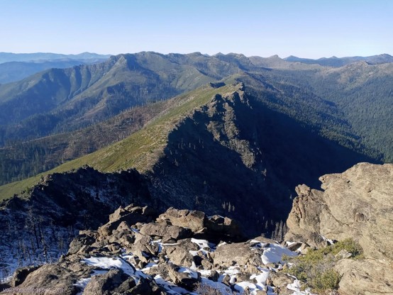 Looking west toward Lonesome Lake, hidden behind the ridge.

A ridge extends away from the viewer, green shrubs rising up the left side, dark rock dropping off the right side. Further on, the steep rock turns to less steep and green. The green on the right drops to a v shaped valley that parallels the ridge.